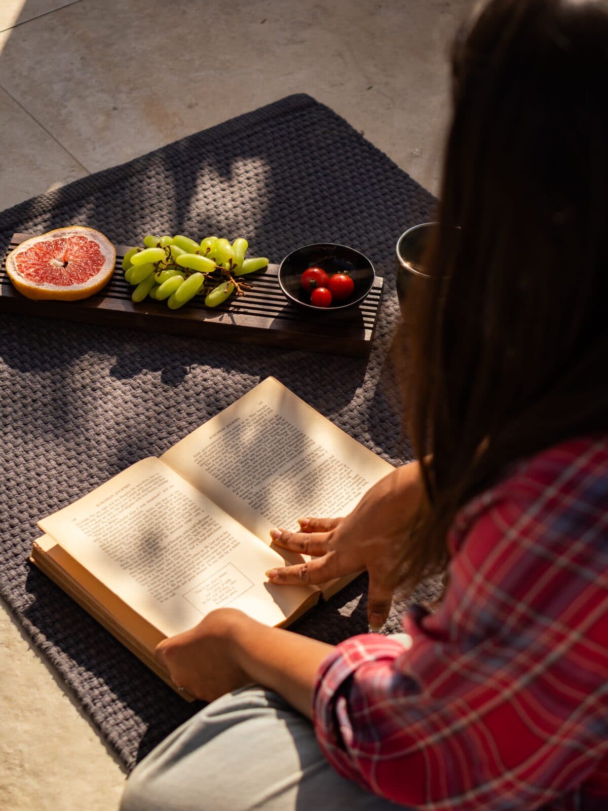A person in a red plaid shirt sits on a dark woven mat outdoors, reading an open book. A fruit tray with a grapefruit slice, green grapes, cherry tomatoes, and a cup is placed nearby on the mat.