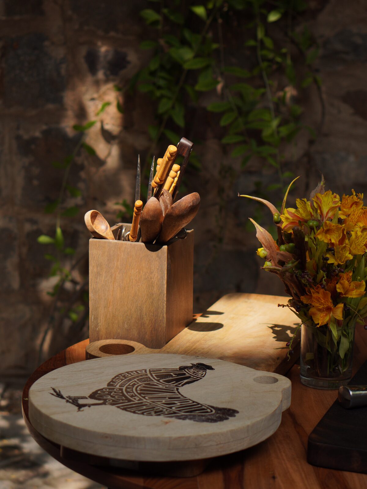 A rustic wooden cutting board with an engraved rooster design sits on a wooden table next to a wooden utensil holder filled with cooking tools, with a vase of yellow flowers in the background.