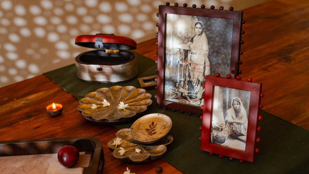 A styled tabletop arrangement featuring two vintage-style wooden photo frames with red trim holding sepia-toned portraits, alongside a small open red-lidded metal box, carved wooden bowls, a lit candle, and a dark green runner on a wooden table.
