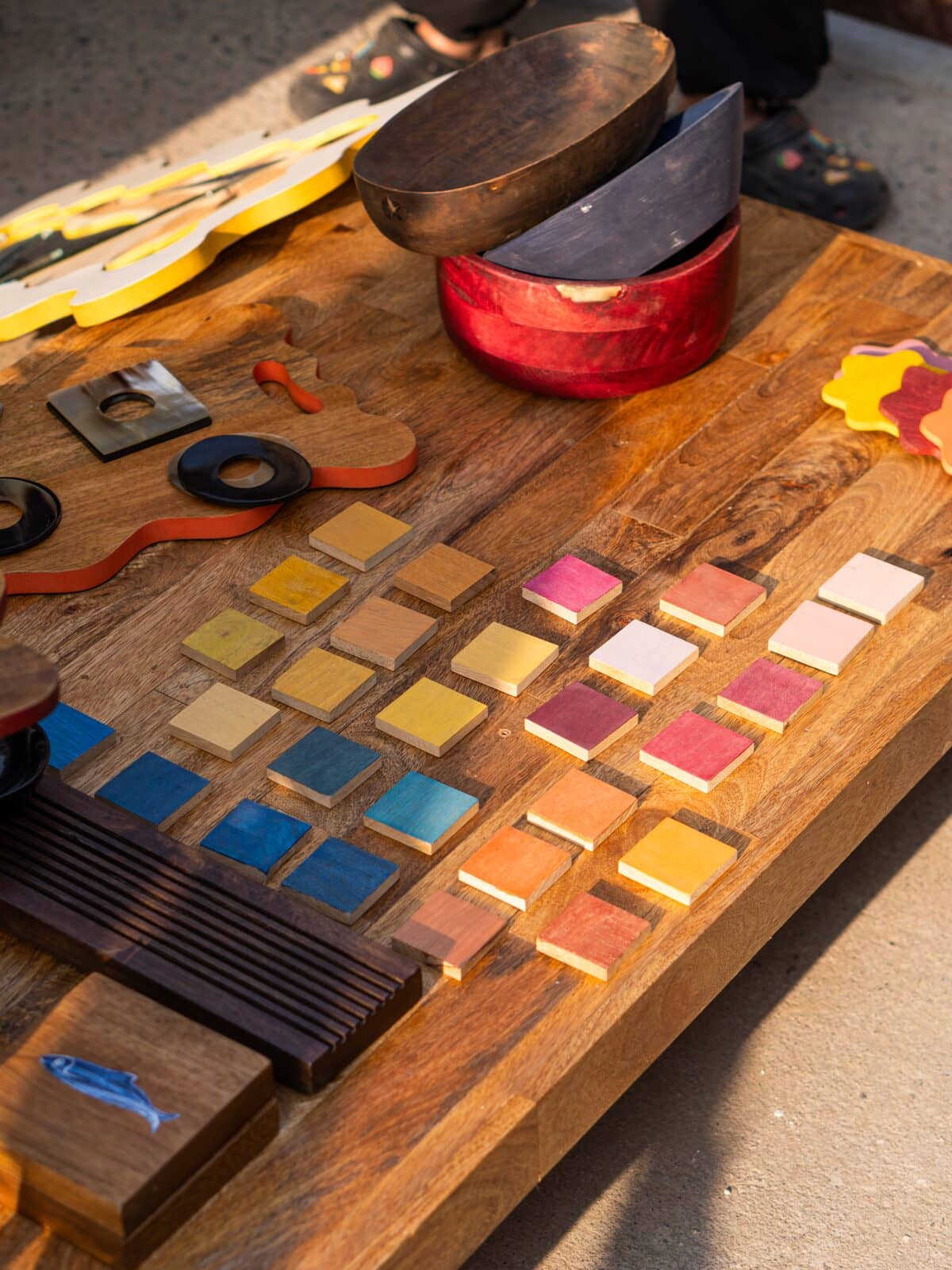A wooden board displaying many small square color chips in different hues, arranged in rows. The board also holds other colorful wooden objects and tools, photographed outdoors on a concrete surface with natural sunlight and shadows.